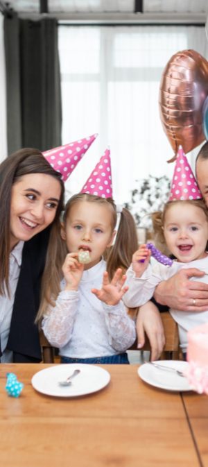 birthday-party-and-family-in-holiday-hats-at-the-table-waving-at-the-camera-pink-cake-and-balloons